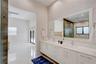 Dual-sink vanity with white cabinetry, light stone-finish countertops, full-width mirror with integrated lighting, and high-gloss tile flooring