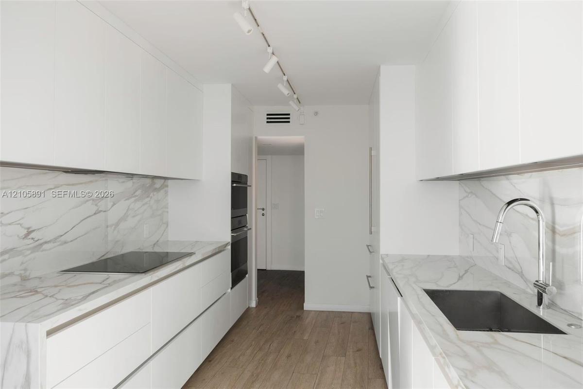 Galley kitchen featuring flat-panel white cabinetry, stone-surface countertops and backsplashes, built-in dual ovens, integrated cooktop, and wood-fin