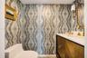 Powder room featuring patterned wallpaper, a wood-finish vanity with sunburst detailing, a stone-composite countertop, an integrated sink, and brass-f