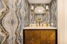 Custom vanity with metallic-textured cabinetry, stone countertop, integrated undermount sink, brass-finish fixtures, and designer wallpaper