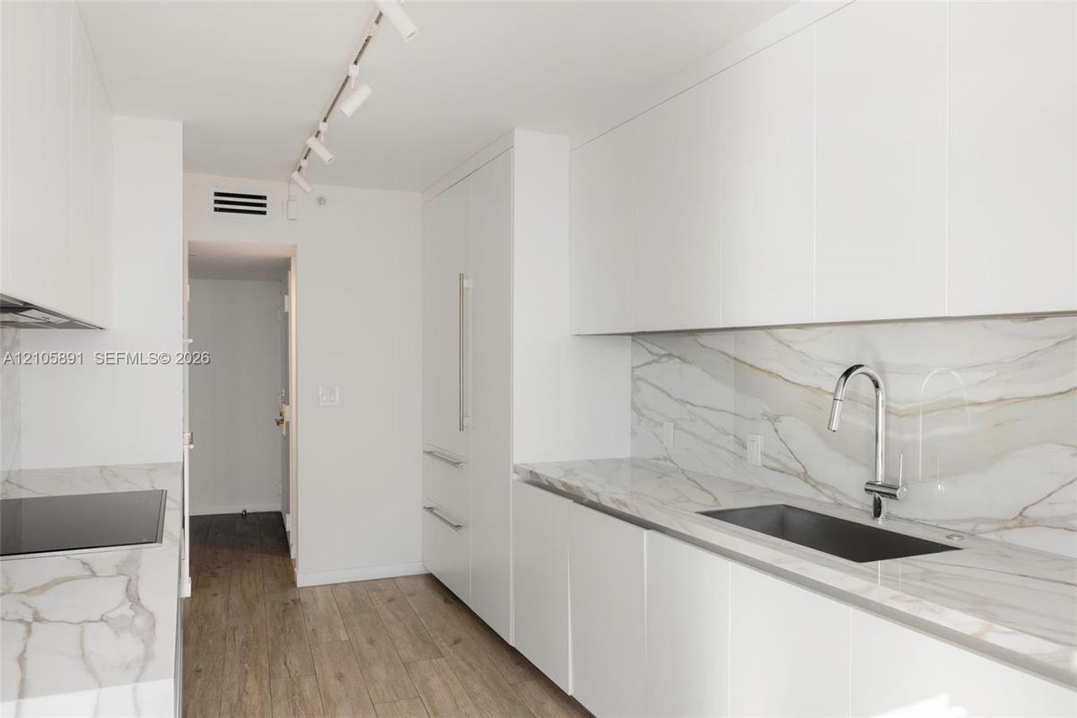 Modern kitchen featuring white flat-panel cabinetry, stone-finish countertops and backsplash, integrated refrigerator, undermount sink with high-arc f