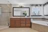 Bathroom featuring wood-finish cabinetry, stone countertop, corner soaking tub with tile surround, glass-enclosed shower, and large framed wall mirror