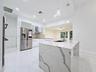 Kitchen featuring a waterfall-edge island with stone-finish countertops, stainless steel appliances, white cabinetry with glass-panel uppers, tile bac