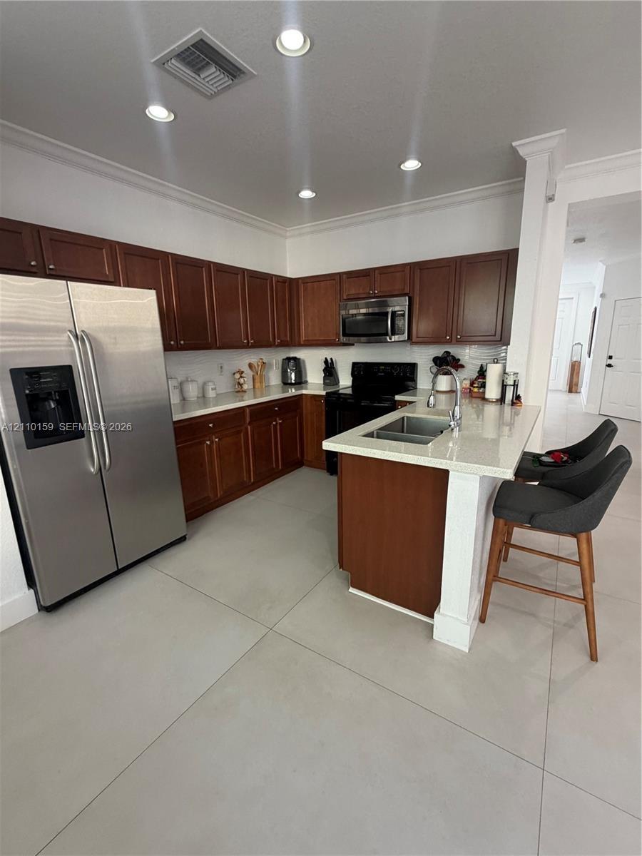Kitchen featuring wood cabinetry, light-toned stone countertops, a center island with a dual-basin stainless steel sink, large-format tile flooring, a