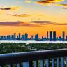 Elevated balcony with a metal railing overlooking a cityscape, waterfront, and lush tree canopy under a vibrant sunset sky
