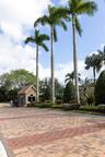 Gated entrance featuring a stone-clad guardhouse, intricate paver driveway, manicured landscaping, and mature palm trees