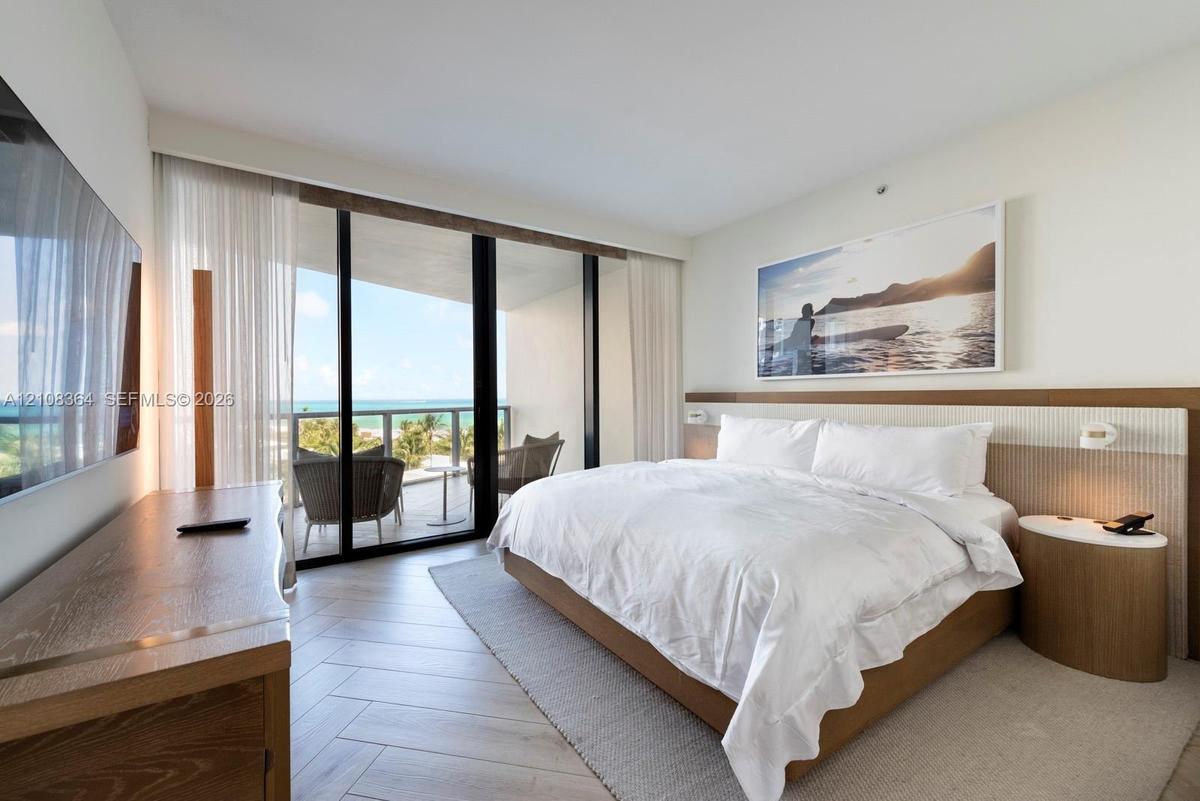 Floor-to-ceiling sliding glass doors, private balcony, wood-finish flooring in a herringbone pattern, and built-in headboard with integrated lighting