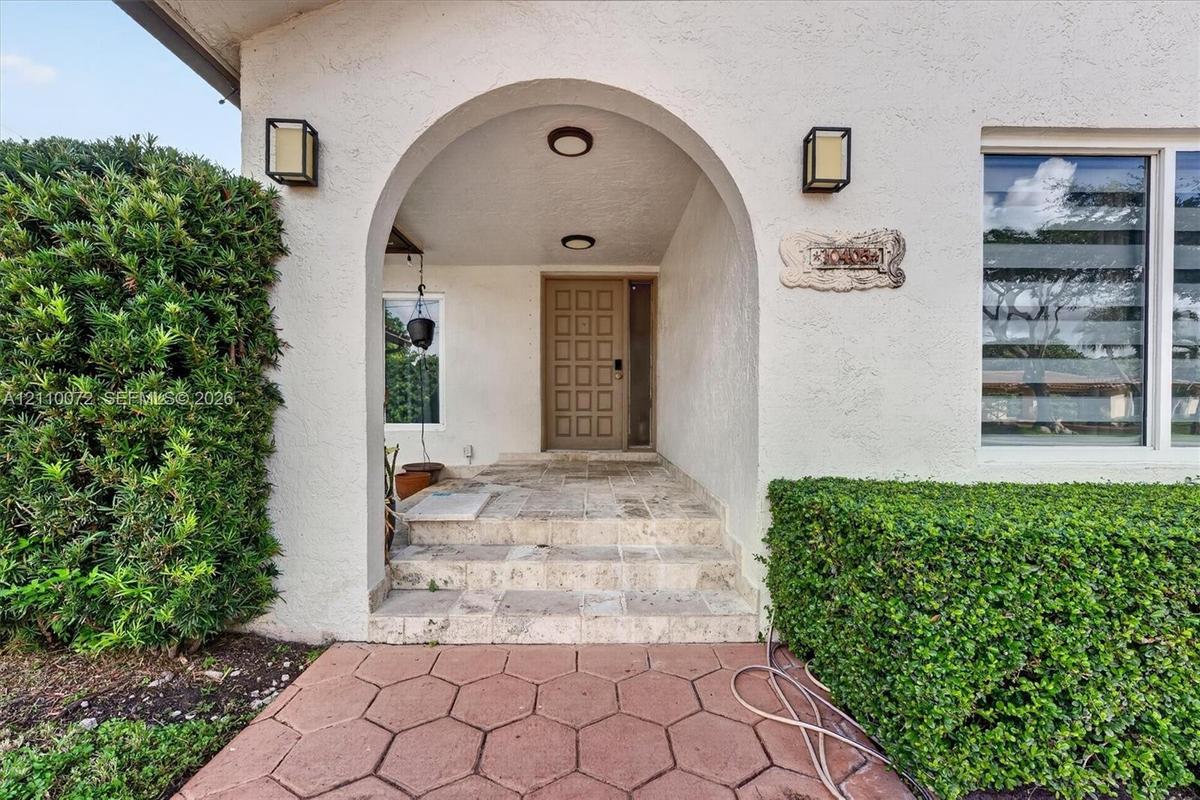 Covered entryway featuring a stone-stepped porch, arched architectural detail, square-paneled front door, and metal wall-mounted light fixtures