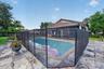 In-ground swimming pool enclosed by a mesh safety fence, surrounded by multi-toned stone paver decking and a covered patio area