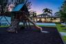 Expansive backyard featuring a built-in wooden play structure with a climbing wall and slide, manicured lawn, established palm trees, and a paved pati