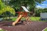 Expansive backyard featuring a wood-frame playground structure, rubberized ground cover, manicured lawn, and mature canopy trees