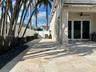 Covered patio featuring stone-tiled flooring and a ceiling fan, complemented by a white vinyl privacy fence and mature palm trees