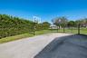 Paved recreational court featuring a built-in basketball hoop, enclosed by chain-link fencing and mature perimeter hedging