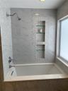 Bathtub surround featuring large-format veined tile, decorative mosaic tile accents, built-in recessed shelving with glass inserts, and polished chrom
