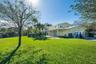 Spacious backyard featuring a manicured lawn, perimeter chain-link fencing, mature trees, a covered patio, and a concrete driveway