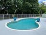 In-ground pool with stone coping, surrounded by a light-toned paved deck and enclosed by white metal fencing