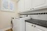 Laundry area featuring white shaker-style cabinetry, dark stone-finish countertops, subway tile backsplash with decorative inset, and plantation shutt