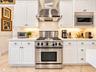 White cabinetry with metallic hardware, stainless steel gas range, professional-grade ventilation hood, built-in stainless steel microwave, and stone 