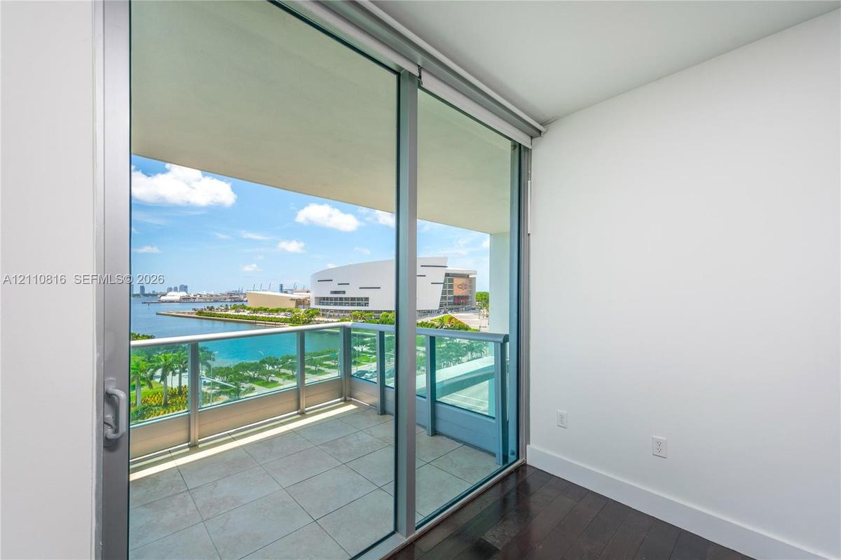 Floor-to-ceiling sliding glass door access to a covered balcony with a metal and glass railing, featuring tile flooring and hardwood-finish interior f