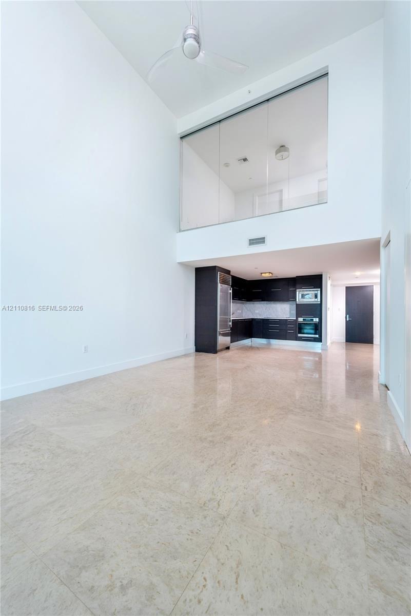 Double-height ceilings featuring an overhead fan, polished stone flooring, dark-toned cabinetry, stainless steel built-in appliances, and a glass-pane