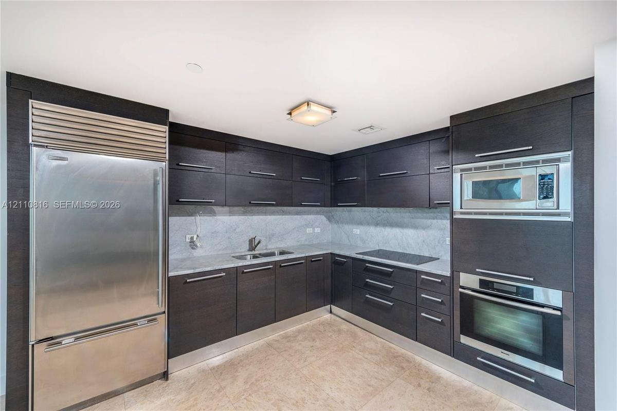 Kitchen featuring dark-toned flat-panel cabinetry, stone countertops, integrated stainless steel refrigerator, built-in microwave, and wall-mounted ov