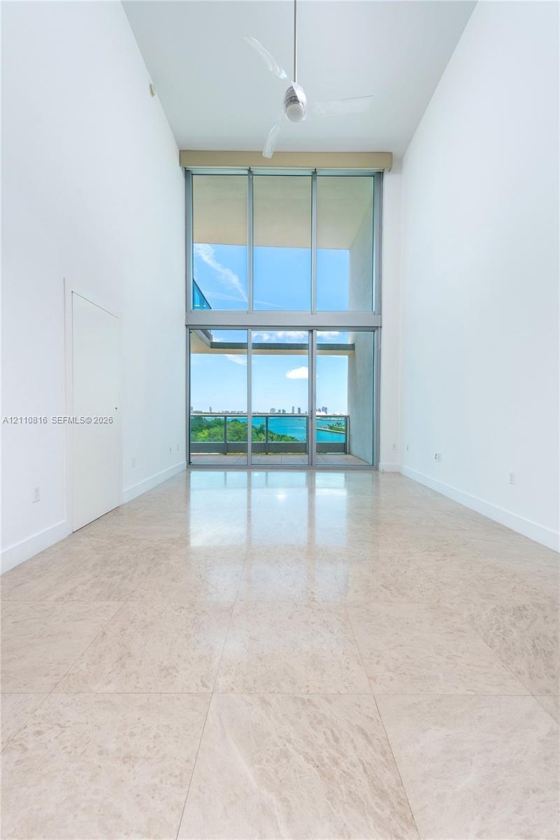 Double-height ceilings, floor-to-ceiling glass wall, polished stone flooring, and ceiling-mounted fan, overlooking water and city skyline
