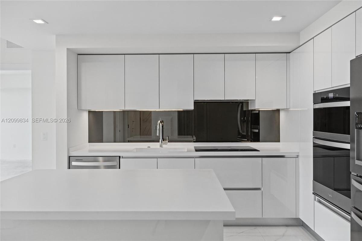 Kitchen featuring high-gloss white cabinetry, white stone countertops, a dark mirrored backsplash, integrated double wall ovens, and recessed lighting