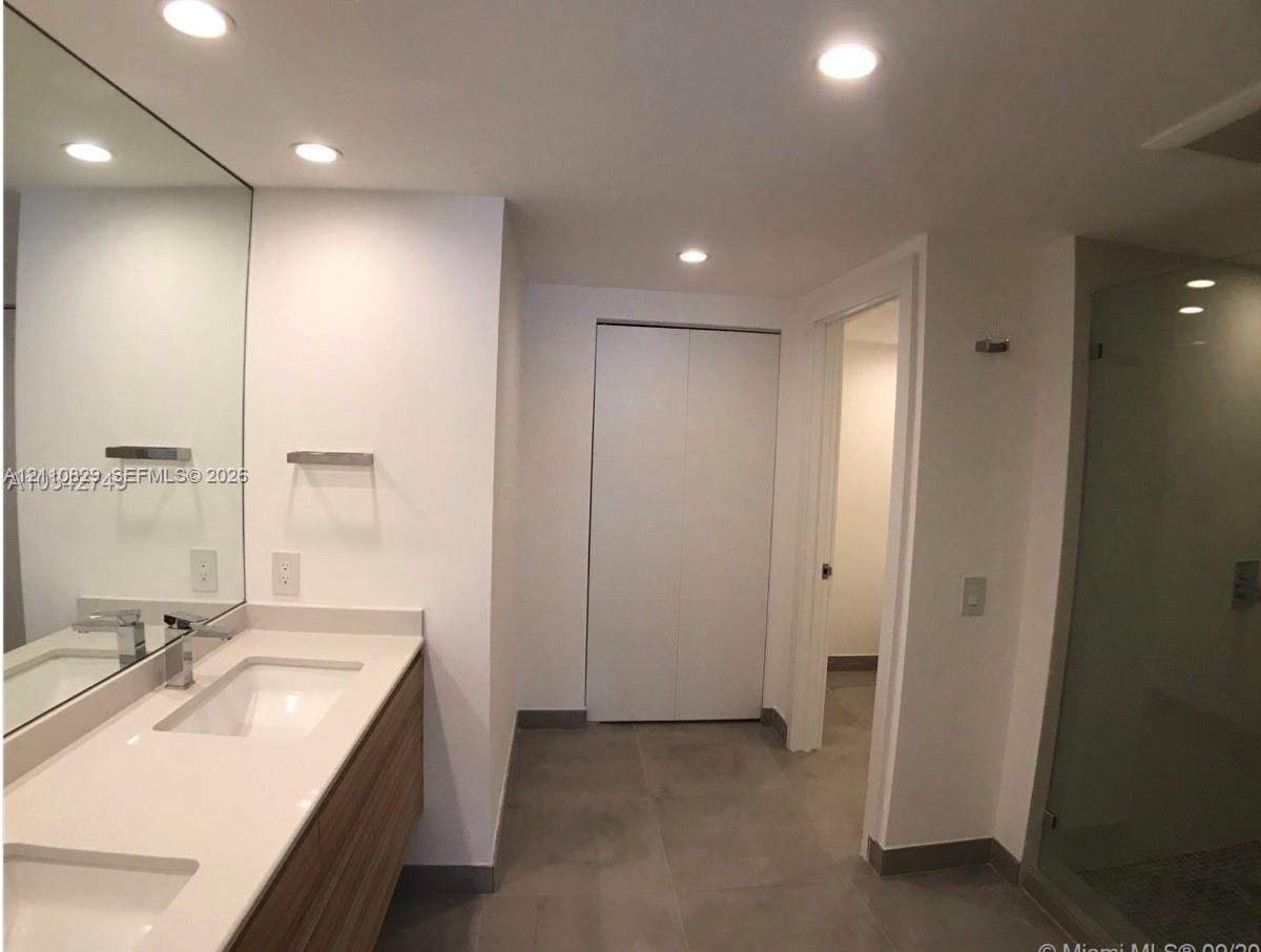 Floating wood-finish double vanity with stone countertop, recessed lighting, expansive wall-to-wall mirror, and glass-enclosed shower with tiled floor