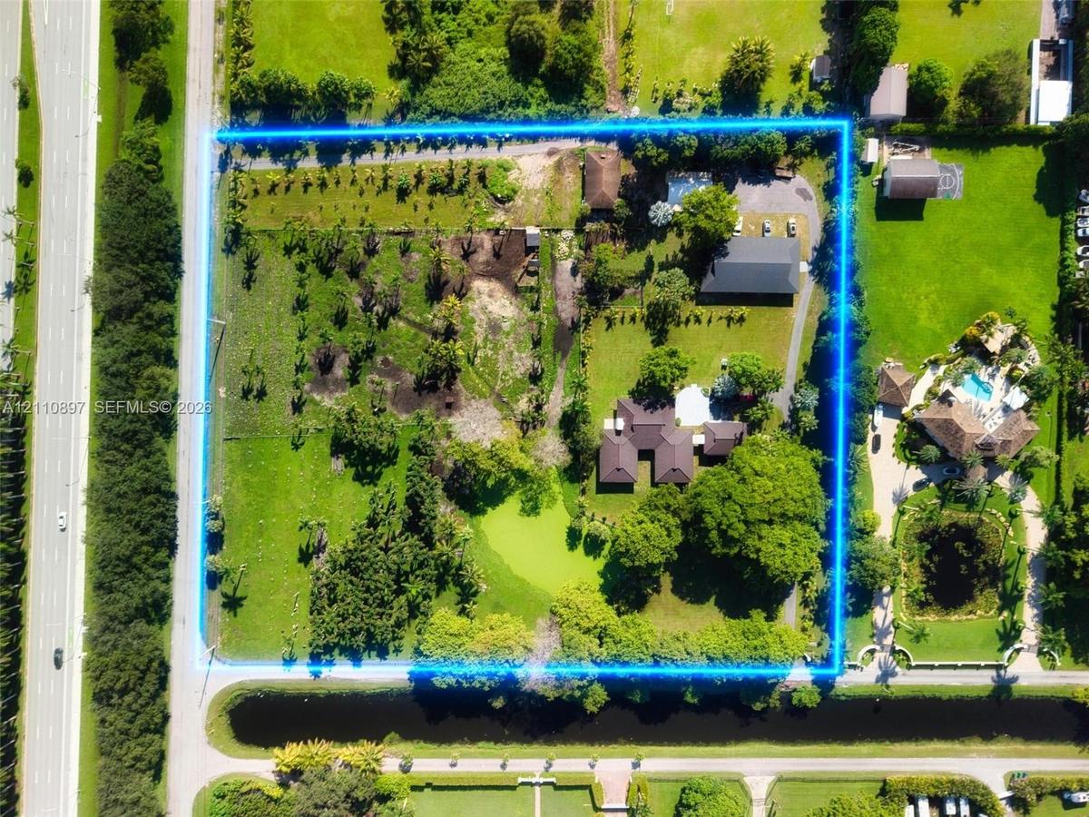 Aerial view of a sprawling rectangular lot featuring expansive grassy acreage, mature trees, a private pond, and multiple structures