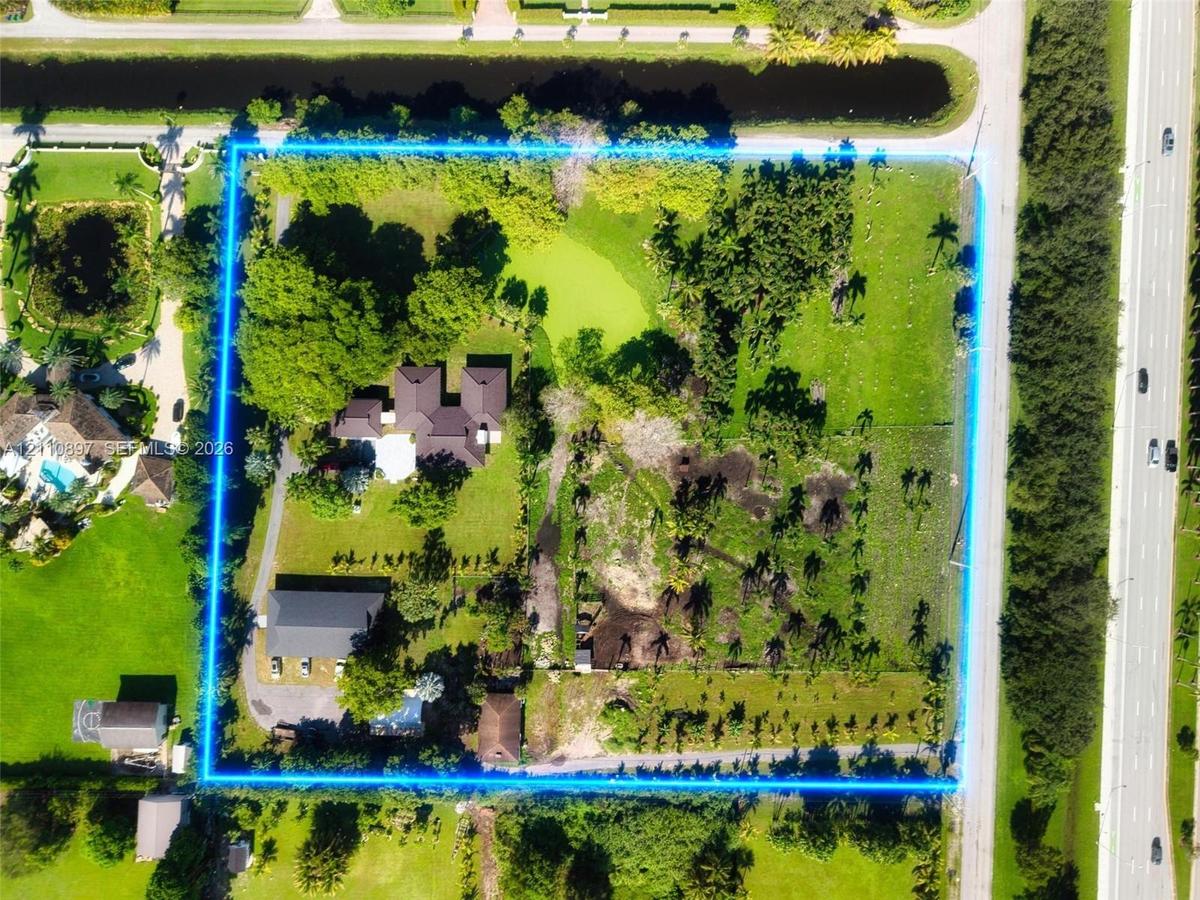 Aerial view of expansive lot featuring a multi-structure property, mature trees, a central pond, and paved driveway access