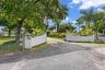 Gated entrance featuring white masonry walls, asphalt driveway, mature trees, and landscaping