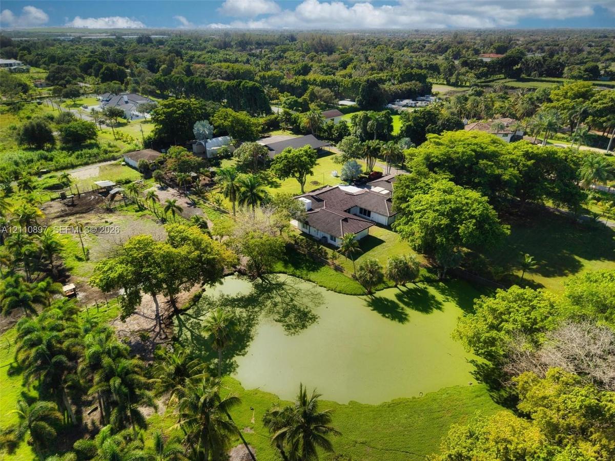 Aerial perspective of a sprawling property featuring a multi-wing residence, a private pond, expansive green space, mature canopy trees, and palm vege