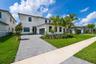 Two-story residence featuring a light-toned exterior, paved driveway, multi-pane windows, attached garage, and landscaped front lawn with palm trees