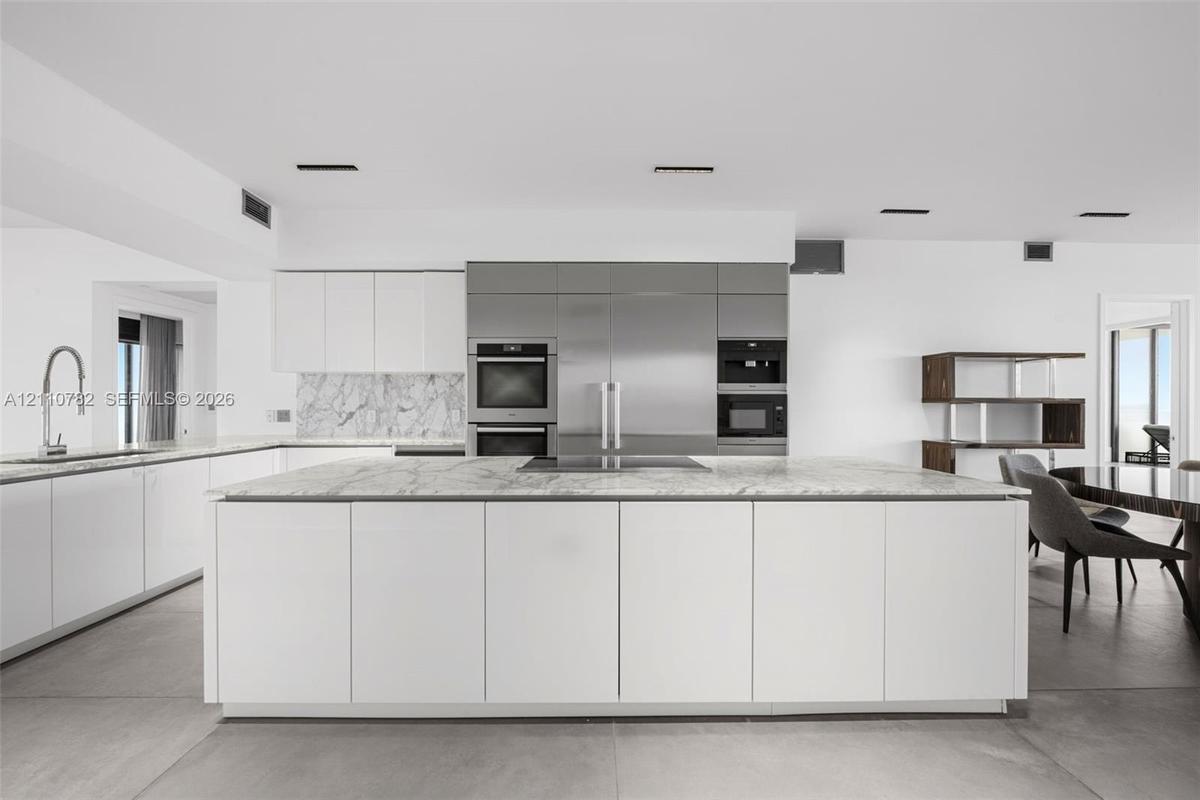 Contemporary kitchen featuring an expansive center island with stone surfacing, stainless steel built-in appliances, flat-panel cabinetry, a stone bac