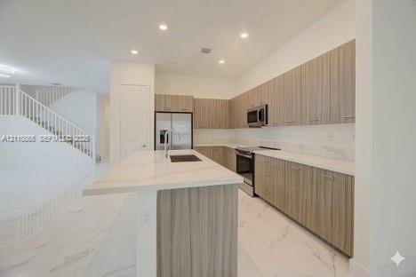 Kitchen featuring flat-panel wood-finish cabinetry, stone-surface countertops, a center island with an integrated sink, stainless steel appliances, an