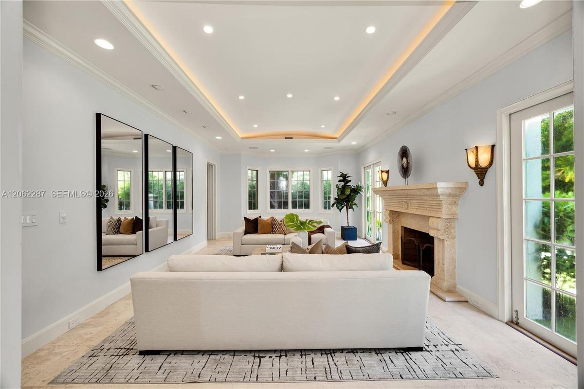 Living area featuring a tray ceiling with recessed lighting, carved stone fireplace mantel, wall-to-wall mirrors, light-toned stone flooring, and mult