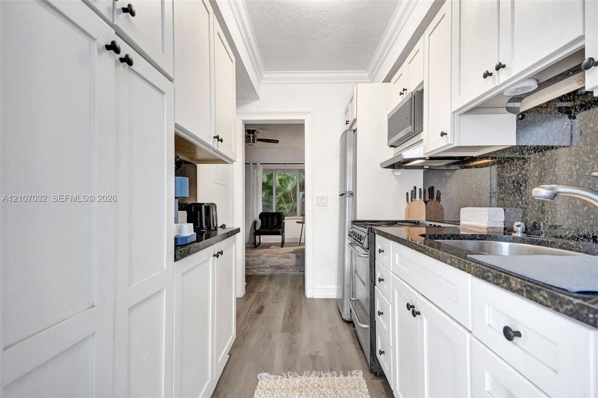 Galley kitchen featuring white shaker-style cabinetry, stone countertops, integrated stainless steel appliances, dark stone backsplash, and wood-finis