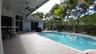 Covered patio featuring a patterned paver floor and ceiling-mounted fan, situated beside a rectangular swimming pool with integrated steps