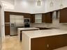 Kitchen featuring flat-panel dark wood cabinetry, waterfall-edge stone countertops, stainless steel appliances, glass-front upper cabinets, and pendan