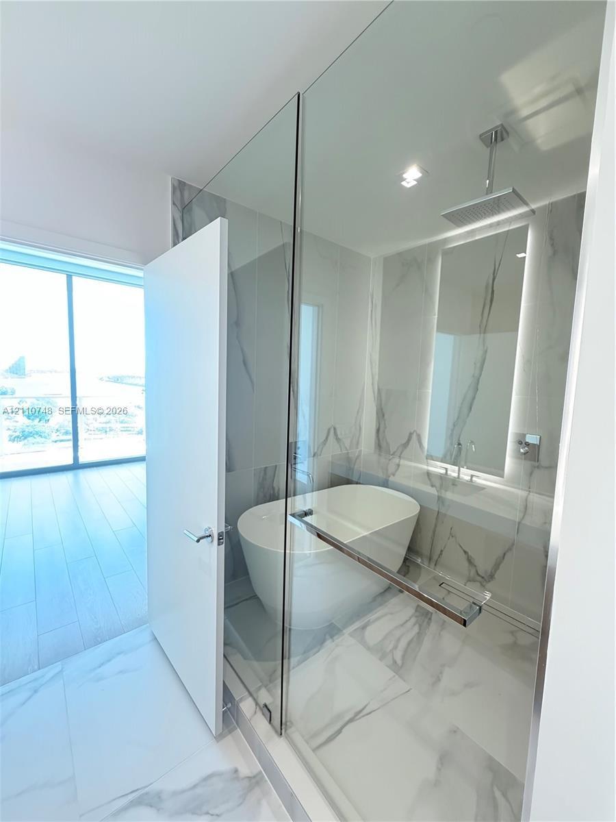 Freestanding soaking tub enclosed in a glass-partitioned wet room featuring floor-to-ceiling stone-look tiling, a ceiling-mounted rainfall showerhead,
