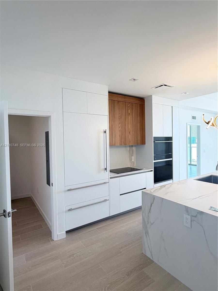Kitchen featuring a stone-surface island, built-in integrated refrigerator, wall-mounted double oven, custom flat-panel cabinetry with wood-finish upp
