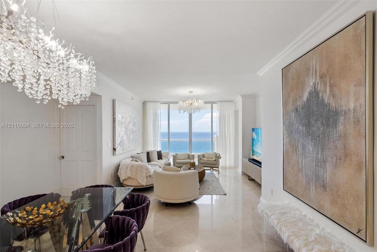 Expansive living area featuring floor-to-ceiling windows with ocean scenery, polished stone flooring, recessed crown molding, and dual crystal chandel
