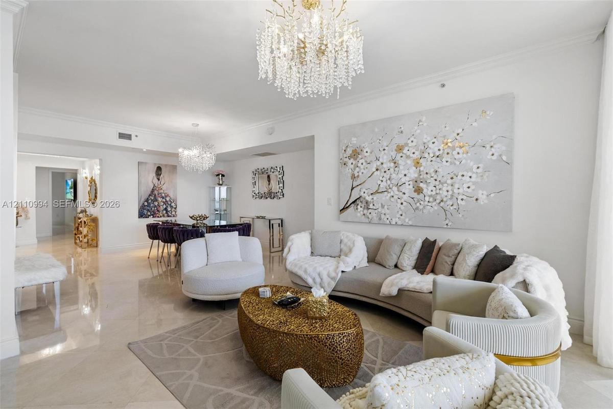 Open-concept interior featuring large-format polished stone flooring, crown molding, and dual ornate crystal chandeliers