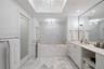 Soaking tub with stone surround, custom tiled accent wall, skylight with recessed tray ceiling, dual crystal chandeliers, and white cabinetry with sto