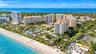 Aerial perspective of oceanfront high-rise structures, featuring expansive sandy coastline, turquoise water, manicured tropical landscaping, and outdo