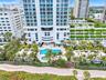 High-rise residential exterior featuring expansive glass balconies, a ground-level swimming pool, a separate spa, landscaped grounds with mature palm 