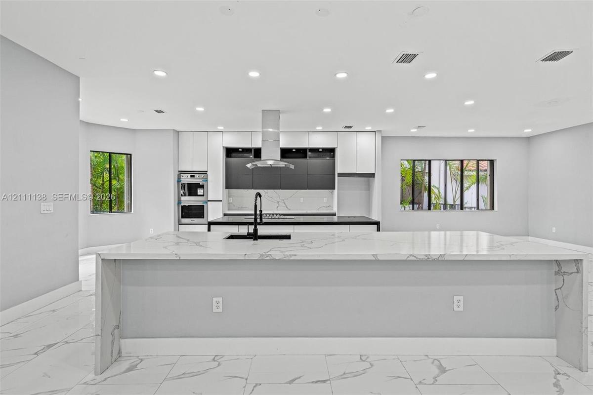 Kitchen featuring a large waterfall-edge island with stone-finish countertops, matte black faucet, integrated stainless steel double oven, two-tone ca