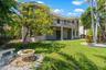 Two-story stucco exterior featuring a curved upper-level balcony with metal railing, classic columns, stone-accented landscaping, and a tiered fountai