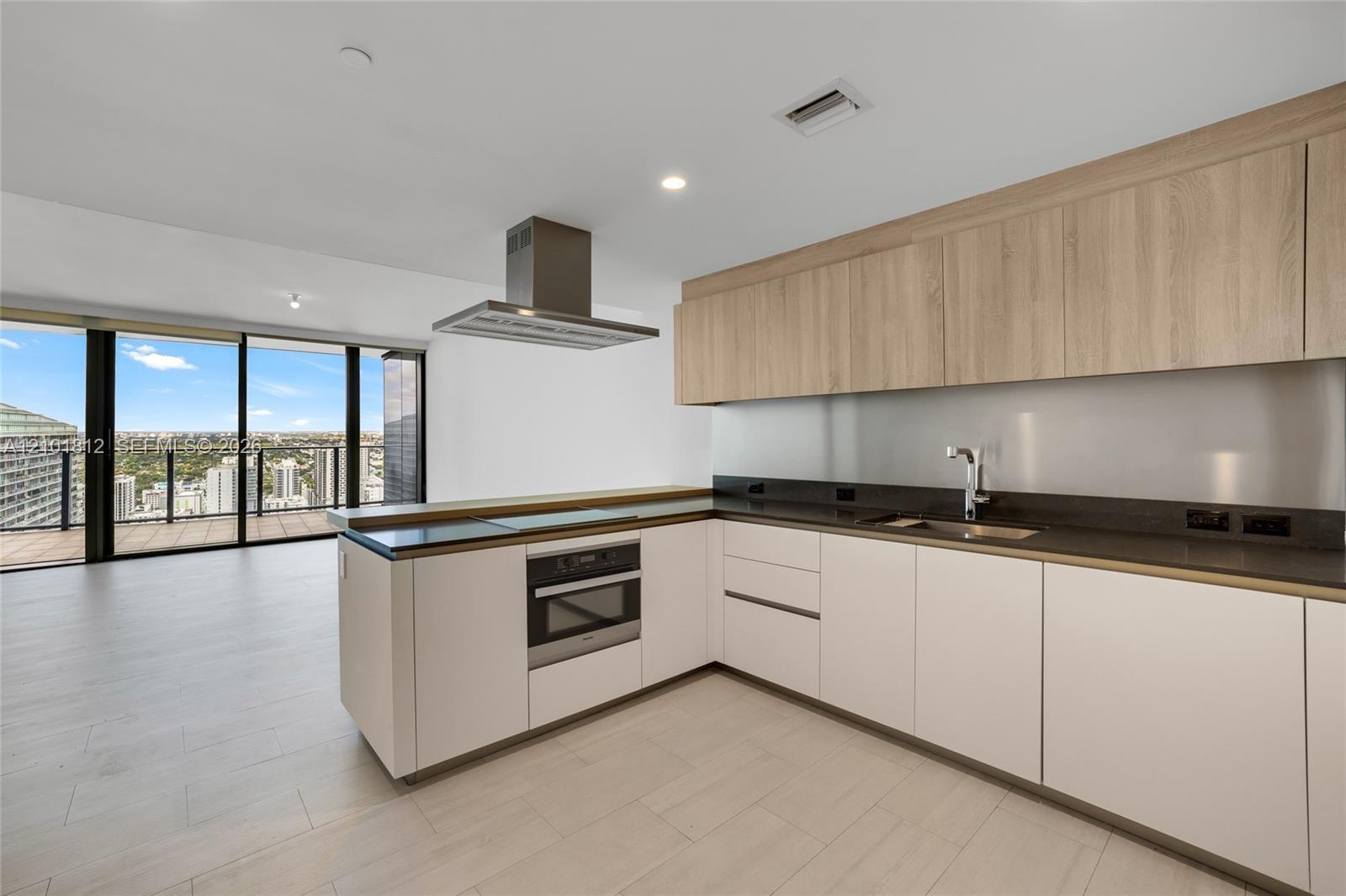 Kitchen featuring flat-panel cabinetry with wood-finish uppers, dark solid-surface countertops, stainless steel wall-mounted range hood, built-in oven