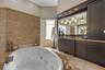 Jetted soaking tub with stone tile surround, custom dark wood cabinetry, integrated lighting, stone-finish countertop, and decorative wall stonework